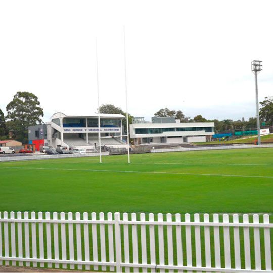 A view across a picket-fenced sporting ground of a grandstand and a modern facilities building.